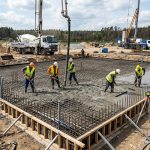 Obraz przedstawia plac budowy w pełnym słońcu, na którym trwają intensywne prace nad wylewaniem betonowej płyty fundamentowej pod budynek mieszkalny lub komercyjny
